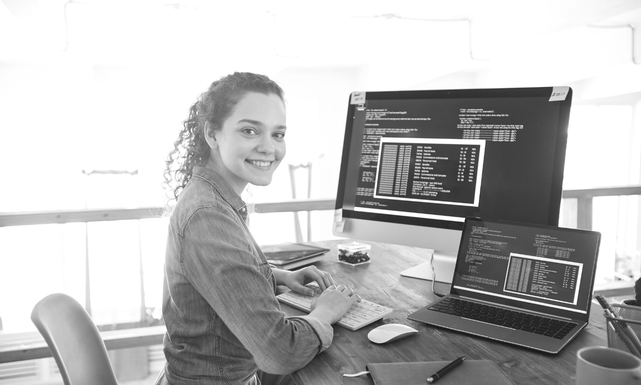 Image of female developer working on two-screen setup and smiling at camera